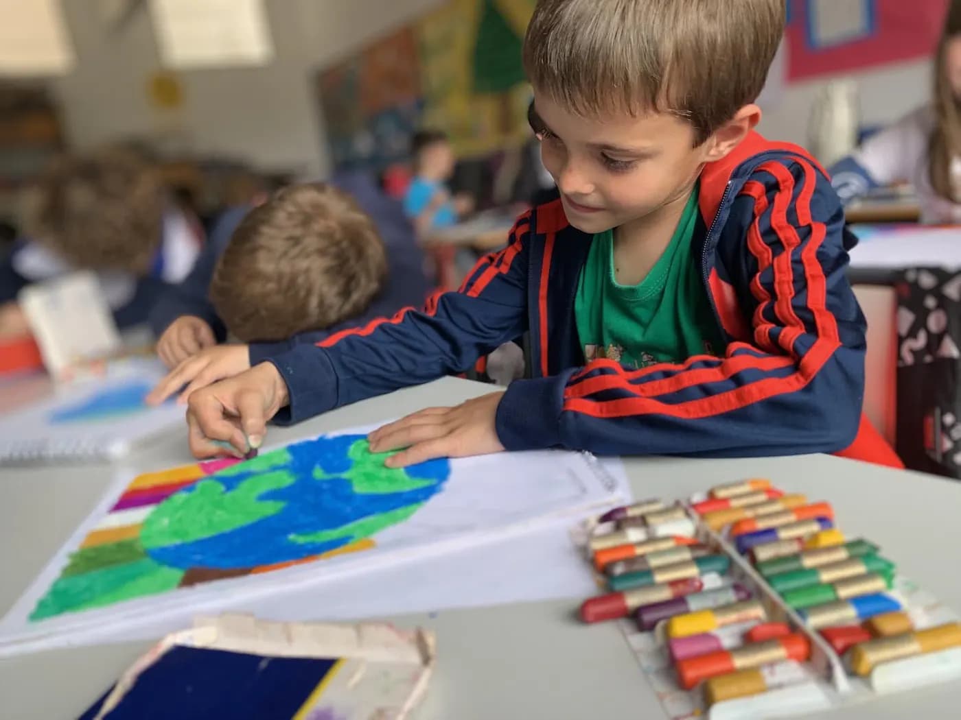 Un alumno de Primaria colorea un mapa del mundo sobre su mesa, con más alumnos trabajando al fondo del aula.