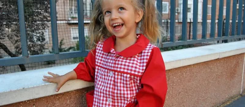 Una niña de Infantil con el babi de cuadros rojos del colegio, sonriendo apoyada en la barandilla del patio.